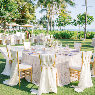Outdoor wedding reception set on a lush lawn with round tables dressed in ivory lace table linens and gold chiavari chairs draped with soft white fabric. Elegant floral centerpieces, glassware, and candles sit beneath string lights and crystal chandeliers, with palm trees and ocean views creating a refined tropical atmosphere.