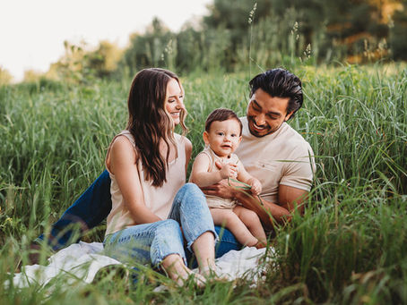 Family of three sitting on a blanket in a lush green field. The parents smile at their cheerful baby. Bright, sunny day.