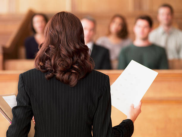 A woman dressed in a pinstripe suit is standing in a courtroom, holding papers and addressing a group of people seated in the jury box. The photo is taken from behind her, showing her back and the attention of the people in the jury box focused on her.