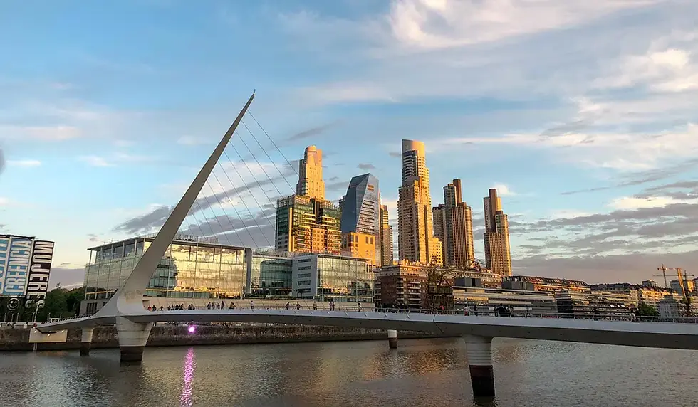 Ponte pedonal moderna sobre o rio, com arranha-céus dourados ao pôr do sol e céu azul com nuvens.