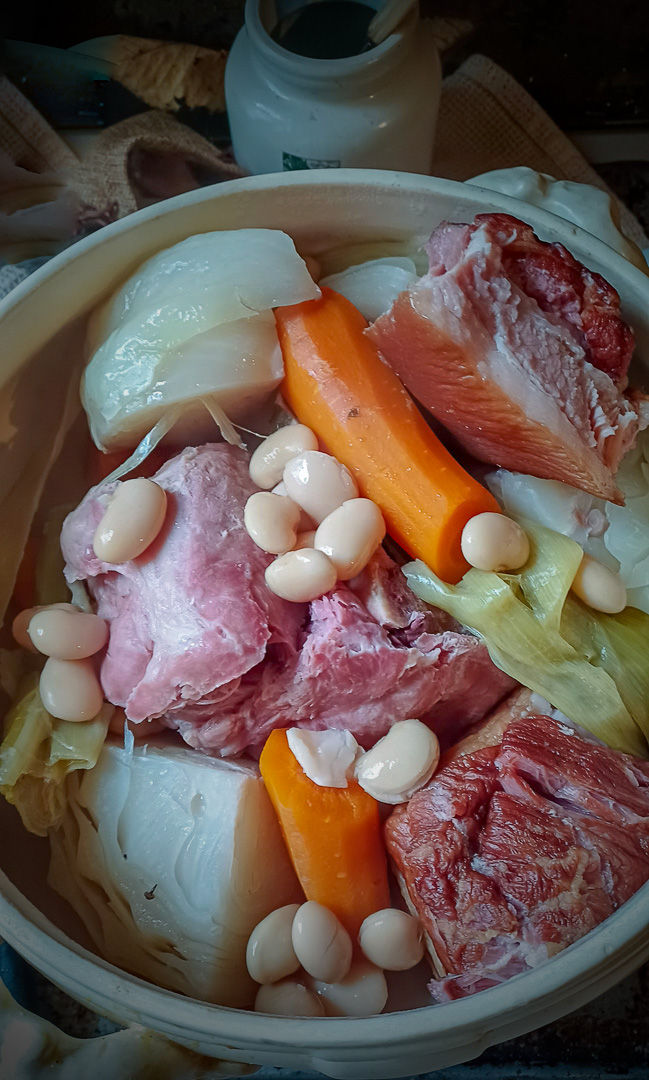L'image montre un plat copieux de potée au chou et petit salé. Le plat comprend des morceaux de viande salée, des carottes, des haricots blancs et du chou, le tout servi dans un pot en céramique rustique. Les couleurs et textures vibrantes des ingrédients rendent le plat appétissant et réconfortant, parfait pour un repas en famille.