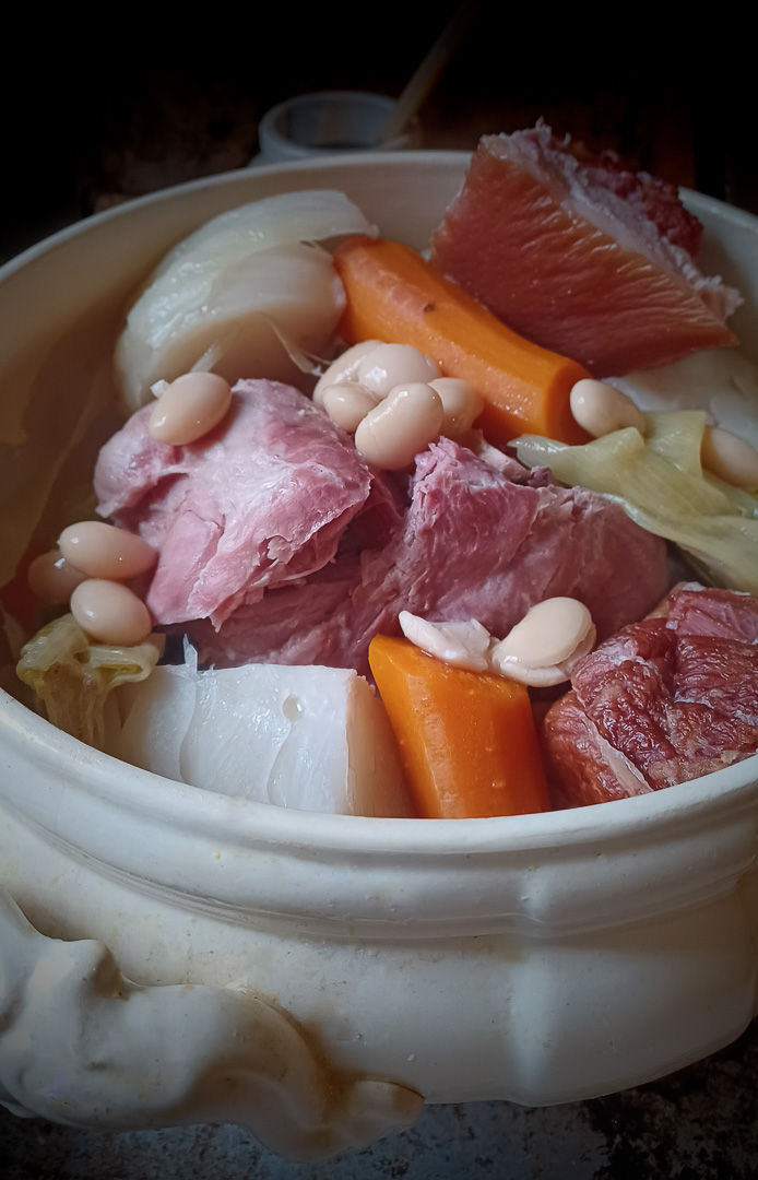 plat copieux de potée au chou et petit salé. Le plat comprend des morceaux de viande salée, des carottes, des haricots blancs et du chou, le tout servi dans un pot en céramique rustique. Les couleurs et textures vibrantes des ingrédients rendent le plat appétissant et réconfortant, parfait pour un repas en famille.