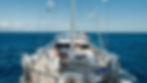 People relaxing on a yacht deck with a clear blue sea and sky. Flags flutter above. The mood is peaceful and leisurely.