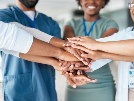 Diverse group of professionals in uniforms stack hands in teamwork gesture, smiling, wearing badges. Bright, collaborative atmosphere.