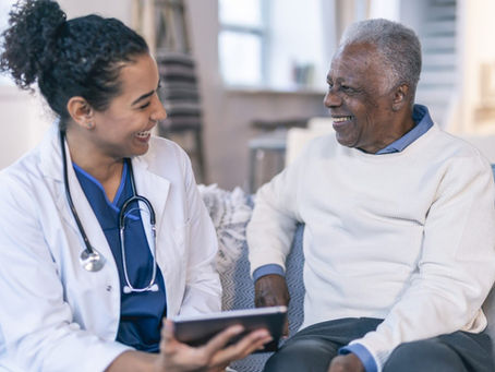 A doctor with a stethoscope and an older man sit on a sofa, smiling and talking. The cozy setting features soft lighting and muted colors.