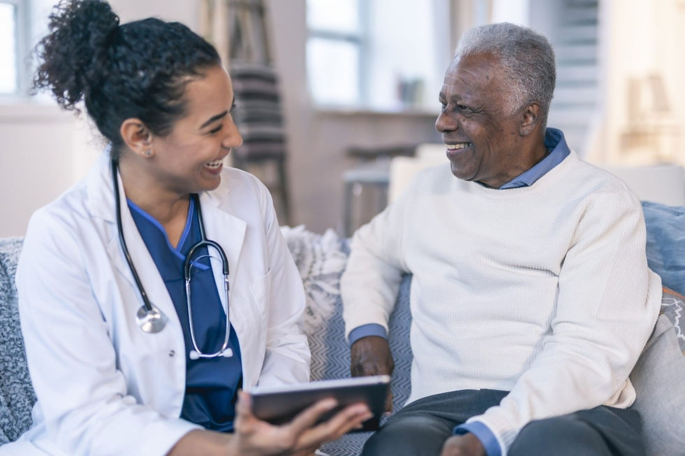 A doctor with a stethoscope and an older man sit on a sofa, smiling and talking. The cozy setting features soft lighting and muted colors.