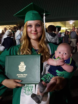 Graduate in a green cap and gown holds a baby and diploma in a crowd. Joyful expression. Text on diploma reads "Slidell High School."