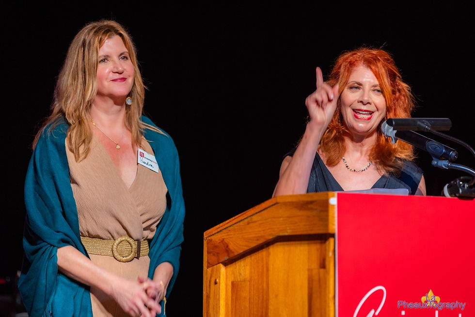 Two women stand at a podium. The woman on the left smiles, wearing a teal shawl. The woman on the right gestures while speaking.