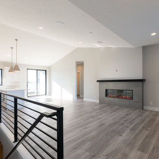 Modern open-concept living space with light gray wood flooring, black metal stair railing, electric fireplace with gray tile surround, white kitchen with large windows, and pendant lighting.