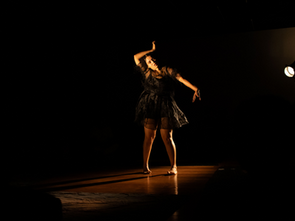 Dançarina de vestido preto posa sob holofote em palco escuro, com braços erguidos e expressão concentrada.