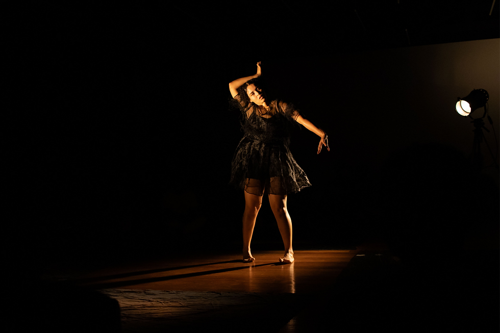 Dançarina de vestido preto posa sob holofote em palco escuro, com braços erguidos e expressão concentrada.