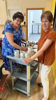 Two elderly ladies, one in a blue dress, one in a dark red top and cream coloured trousers serving hot drinks.