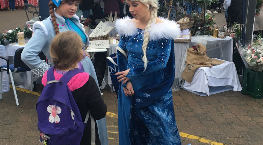 A kid with a pink backpack looking at 2 adults dressed as princesses from a Disney movie called Frozen.