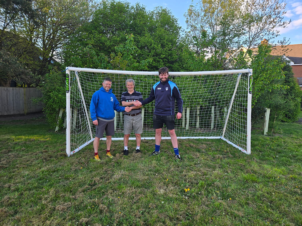 Three men stand smiling in front of a soccer goal, shaking hands. They're in casual sportswear, with trees in the background.