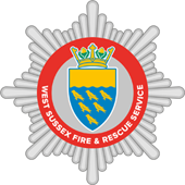 Badge of West Sussex Fire & Rescue Service with a red circular border, crown, blue shield, gold fish design, and radial white background.
