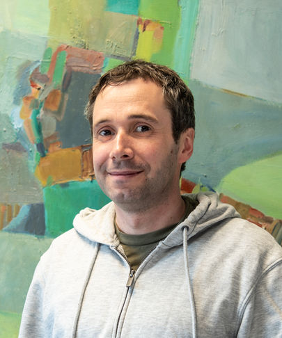 Man in a grey hoodie smiles in front of a colorful abstract painting with green, blue, and orange tones.