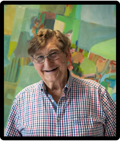 Smiling elderly man in glasses and a red-blue checkered shirt stands before a colorful abstract painting backdrop.