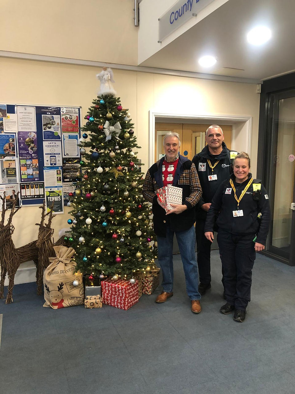 2 men and a woman standing by a Christmas tree, holding gifts and smiling.