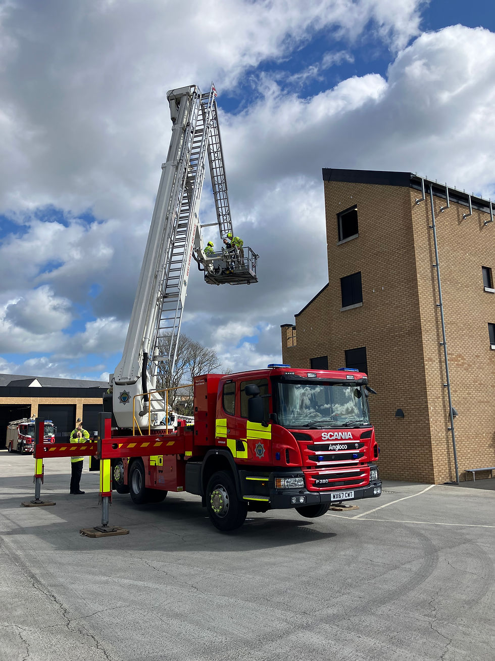 A fire engine with an extended aerial ladder raised high beside a training building, with firefighters standing on the platform.