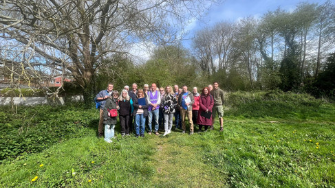 A group of people standing on a grassy path in a green open space, posing for a group photo beneath leafless trees with houses visible in the background on a bright day.