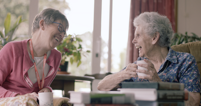 Two women laughing and chatting while holding mugs. They sit in a cozy room with books and plants in the background. Bright ambiance.