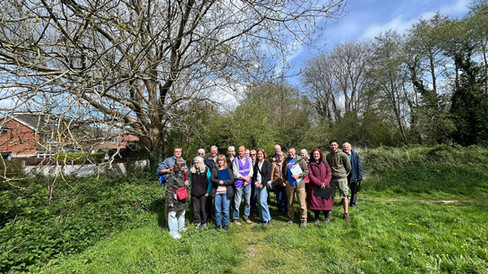 A group of people standing on a grassy path in a green open space, posing for a group photo beneath leafless trees with houses visible in the background on a bright day.