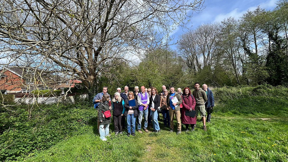 A larger group of people gathered closely together on a grassy clearing, facing the camera for a group photograph with trees and hedges surrounding the area.