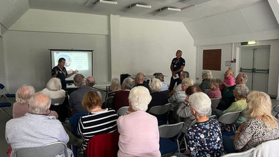A person presents to a seated audience in a bright room. A screen displays "Southwater Neighbourhood Wardens." Attentive and engaged mood.