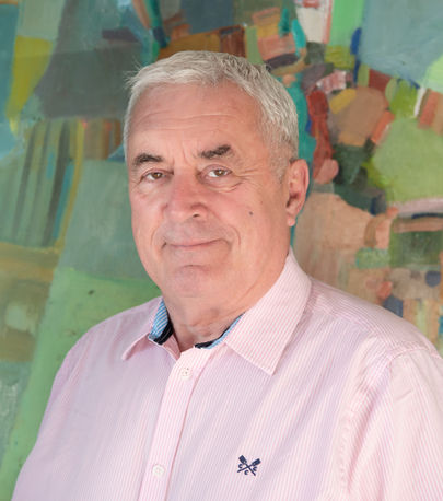 Elderly man in a pink striped shirt smiling in front of a colorful abstract painting. The mood is calm and positive.