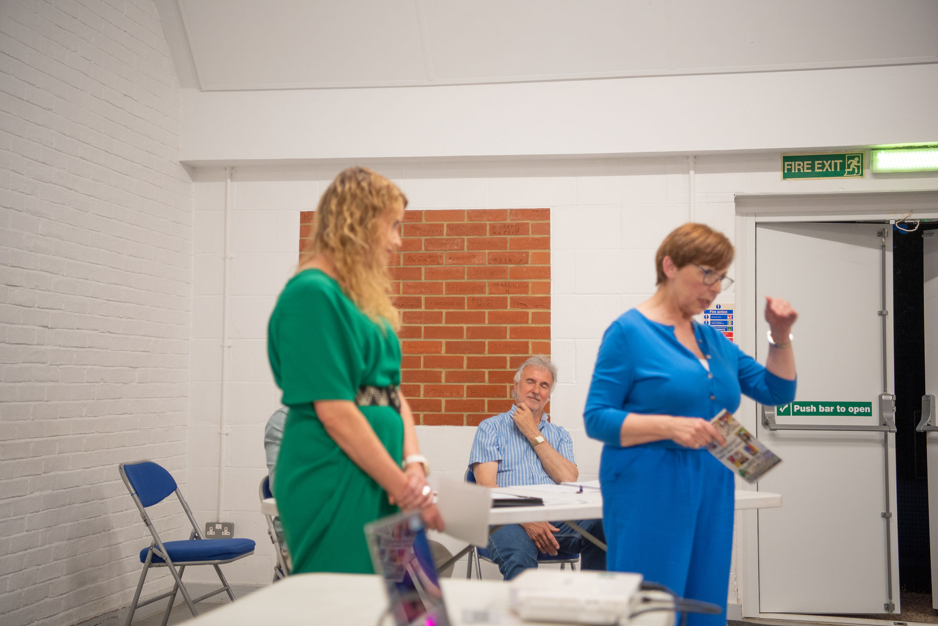 A lady in a green dress and another one in a blue dress are presenting to a group.