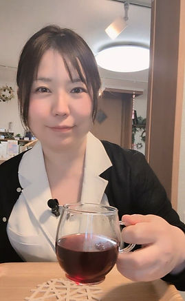 千葉県旭市Healing Salon Torusのセラピストたみが、  サロン内でハーブティーを手に微笑んでいる写真。白と黒いサロンユニフォームを着用。背景にはナチュラルなインテリアとグリーンが見える。ご来店時にはウェルカムハーブティーをご用意しています。