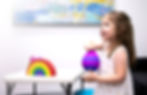 A joyful little girl plays with a purple and pink stress ball, looking engaged and happy