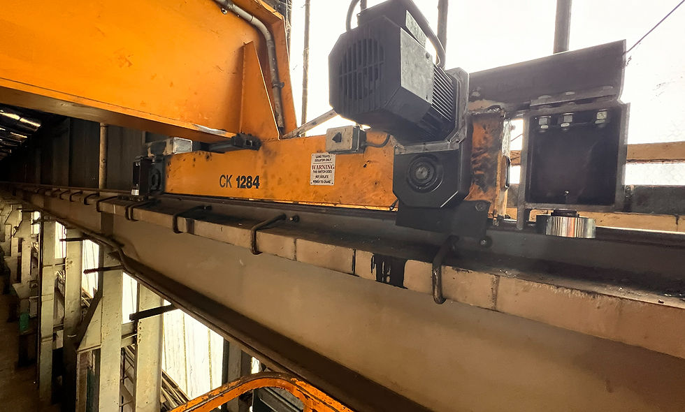 Large crane lifts object at industrial site with gantry structure, pipes, and safety barriers.