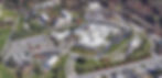 Aerial view of a large hospital complex with multiple buildings, parking lots, and surrounding greenery. Roads and empty lots are visible.