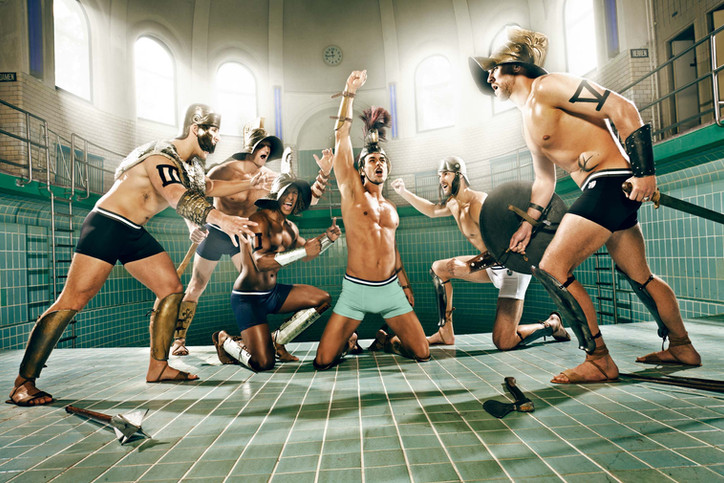 Marcus Schenkenberg and members of the German national water polo team in gladiator-inspired swimwear, performing a dramatic scene in a historic indoor pool – QVC campaign photographed by Erik Bont