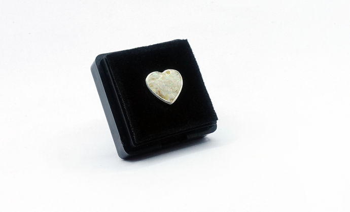 A sterling silver charm in the shape of a heart in a black jewellery box. The heart contains cremated ashes and white snow opal in clear resin.