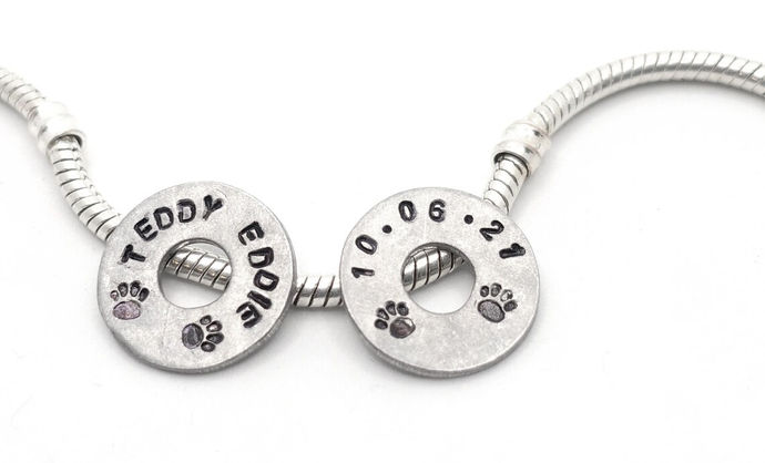 A silver charm bracelet on a white background, with two round aluminium tags with circular central holes resting against it. The tags are hand stamped with words and paw prints. The left tag spells the name 'TEDDY EDDIE' and the right tag has the date '10.06.21'. The indentations made by the stamps are coloured in black.