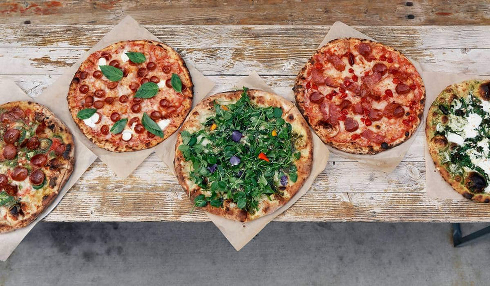 Five assorted pizzas on parchment paper atop a rustic wooden table, topped with pepperoni, basil, greens, and cheese.