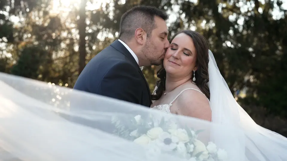 Man kissing woman's cheek; wedding day, happy couple, veil, outdoor setting.