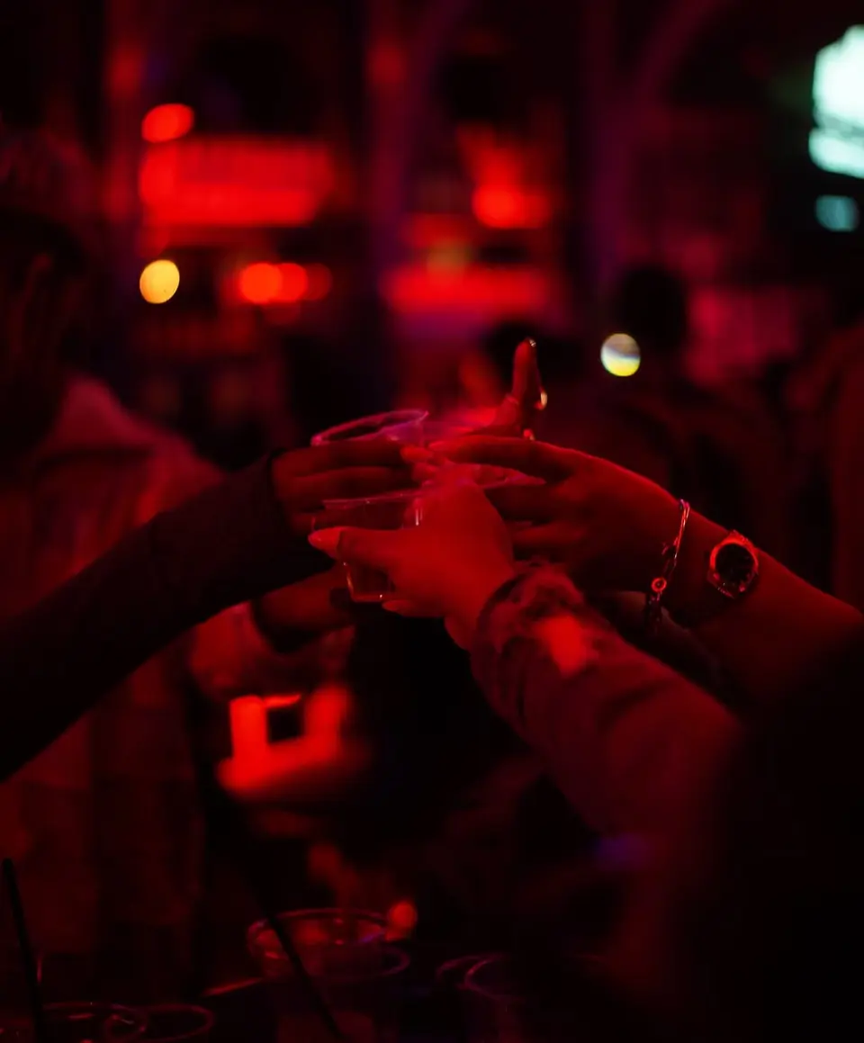 Two hands toasting with drinks in a dark bar, red lighting.