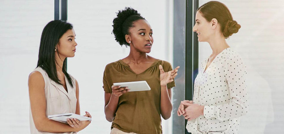 Three women in a bright office talk in a small meeting, two holding notebooks, one gesturing as they discuss.