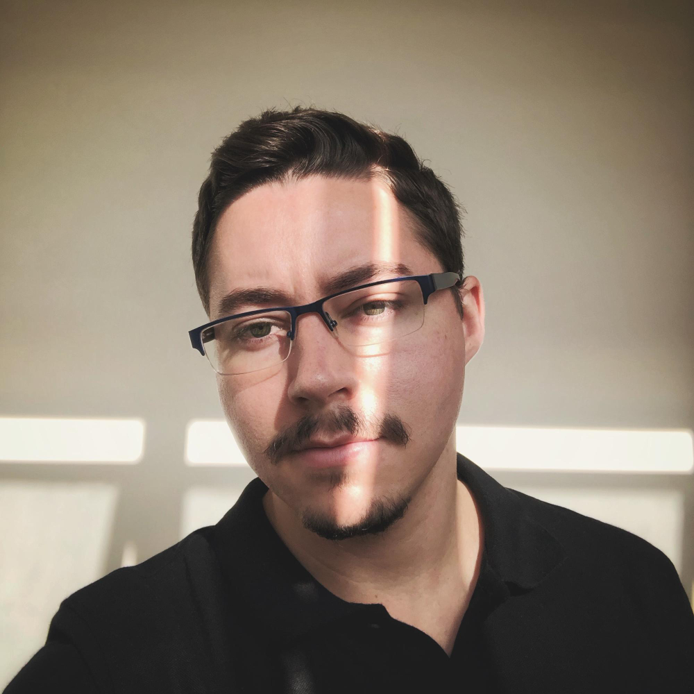 Man with glasses and mustache, facing the camera with dramatic lighting.