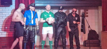 Men in leather and rugby attire on a stage gallery.
