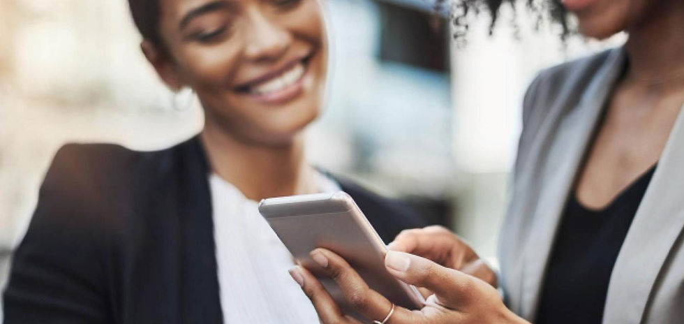 Two smiling women in blazers look at a smartphone outdoors, sharing a cheerful moment.