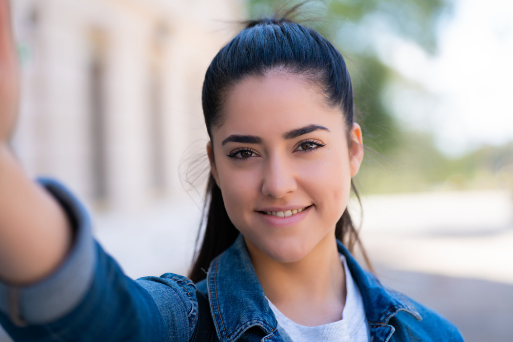 Young woman smiles and takes a selfie wearing a denim jacket outdoors.