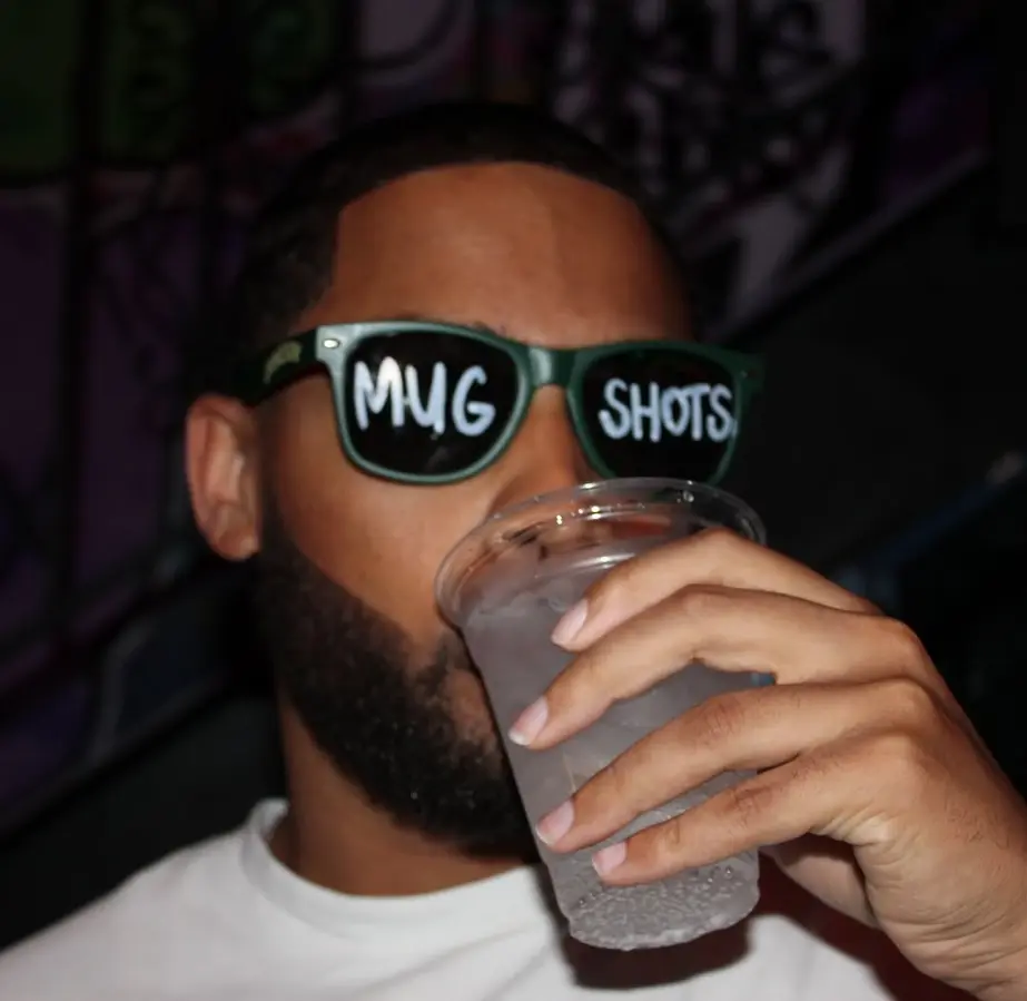 Man wearing sunglasses with "MUG SHOTS" text drinking a beverage dev-mugshots