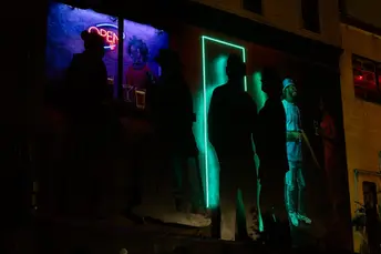 Silhouetted people stand outside a bar with neon signs and inviting light in Baltimore.