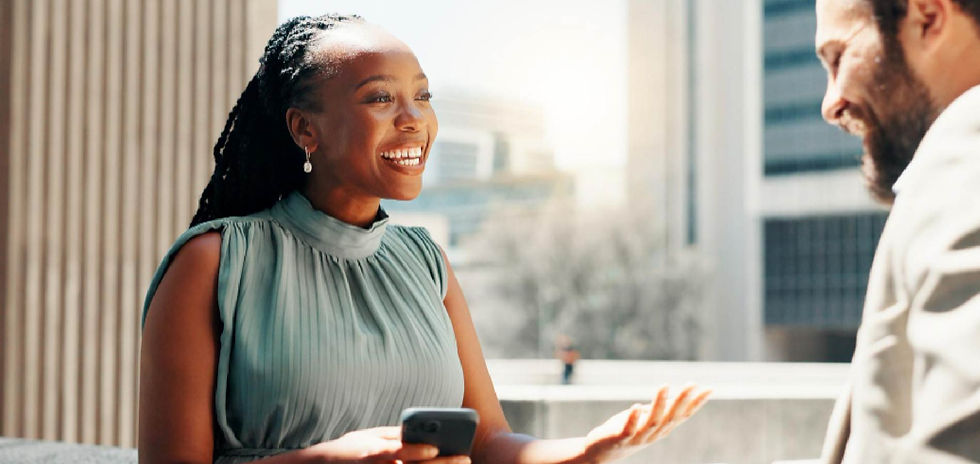 Smiling woman in a teal dress talks with a bearded man on a sunny balcony, holding a phone, with city buildings behind.