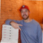 Man smiles, poses with peace sign and pizza boxes, brick wall background.