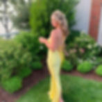 Woman wearing yellow dress standing outside, holding a glass, with green bushes